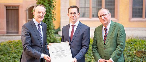 Der Dekan, Prof. Dr. Joachim Suerbaum (links) überreicht die Urkunde zum Abschluss des Habilitationsverfahrens an Dr. David Kuch (Mitte) in Anwesenheit von Prof. Dr. Horst Dreier (rechts).