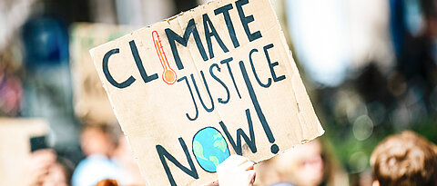 Ein Schild im Rahmen einer Demonstration mit der Aufschrift "CLIMATE JUSTICE NOW!".